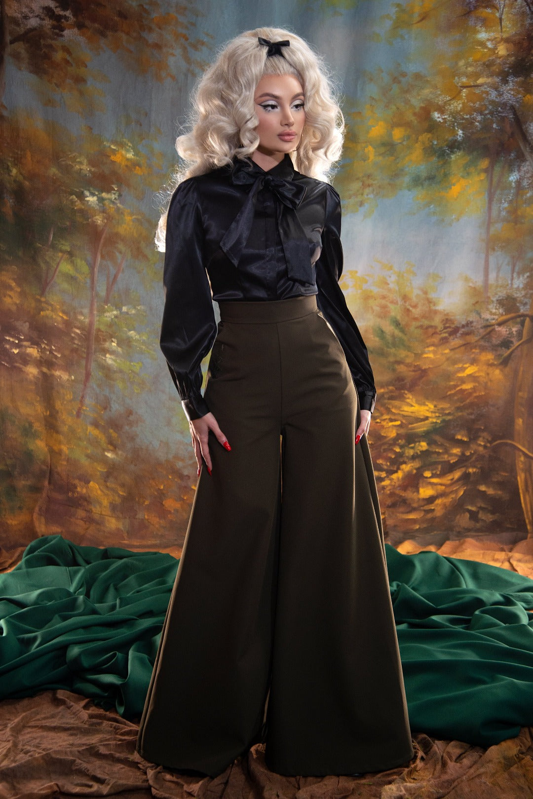 A blond pinup model posing against a yellow and blue painted background while wearing the Angela Blouse in black and brown wide-leg trousers.