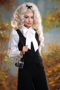 A blond pinup model posing against a yellow and blue painted background while wearing the Angela Blouse in white with a black vest on top and black trousers holding an old camera on her hand.